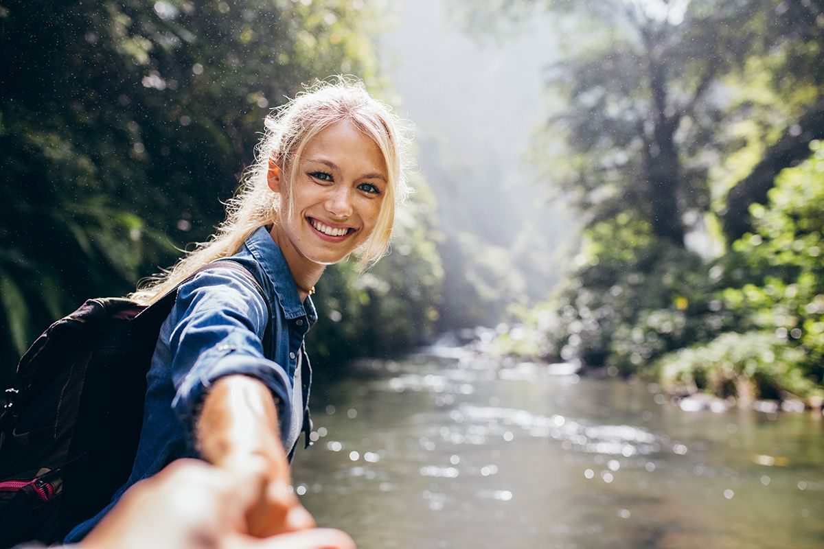 Na zdjęciu jest uśmiechnięta kobieta, która jest nad wodą w lesie i trzyma za rękę mężczyznę. 